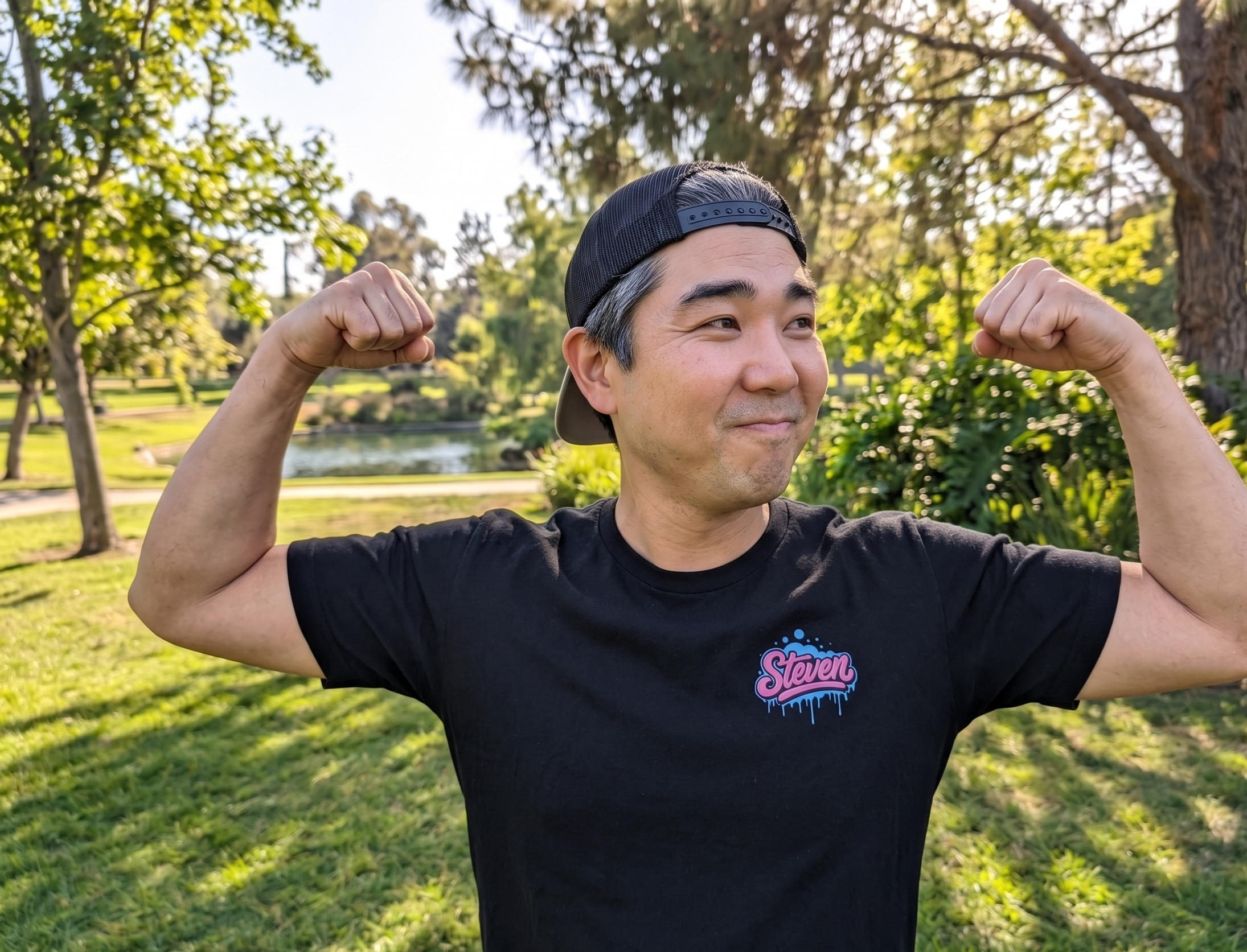 Man flexing muscles outdoors in a park wearing a black t-shirt with a logo.